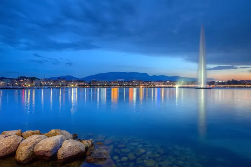 The Jet d'Eau fountain on Lake Geneva at dusk, with city lights and mountains